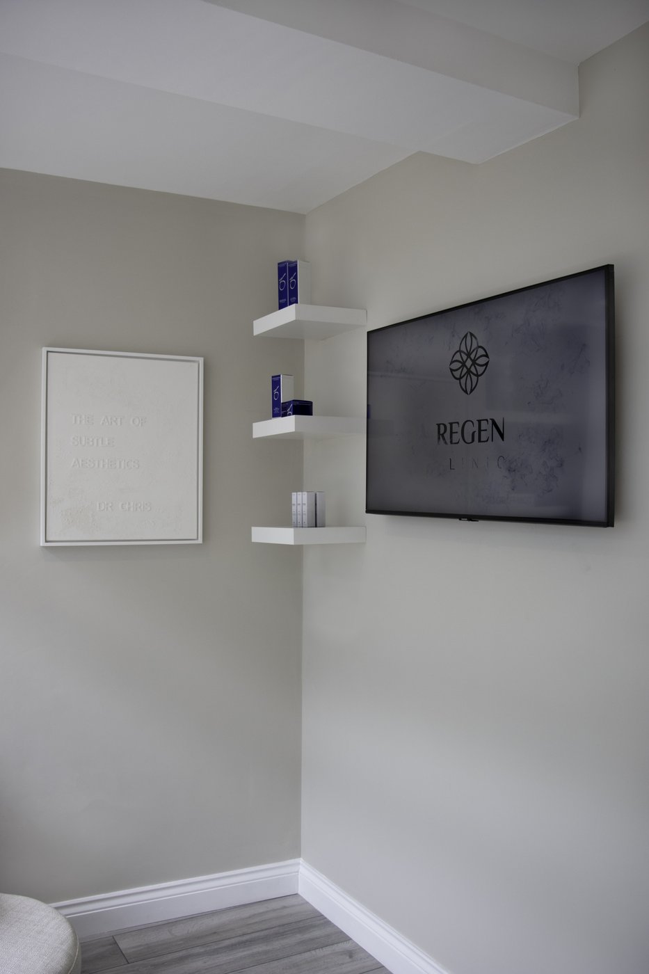 REGEN Clinic interior detail: a textured canvas reading 'The Art of Subtle Aesthetics — Dr Chris' on the wall, white floating shelves with ZO Skin Health products, and the REGEN Clinic logo displayed on a dark screen.