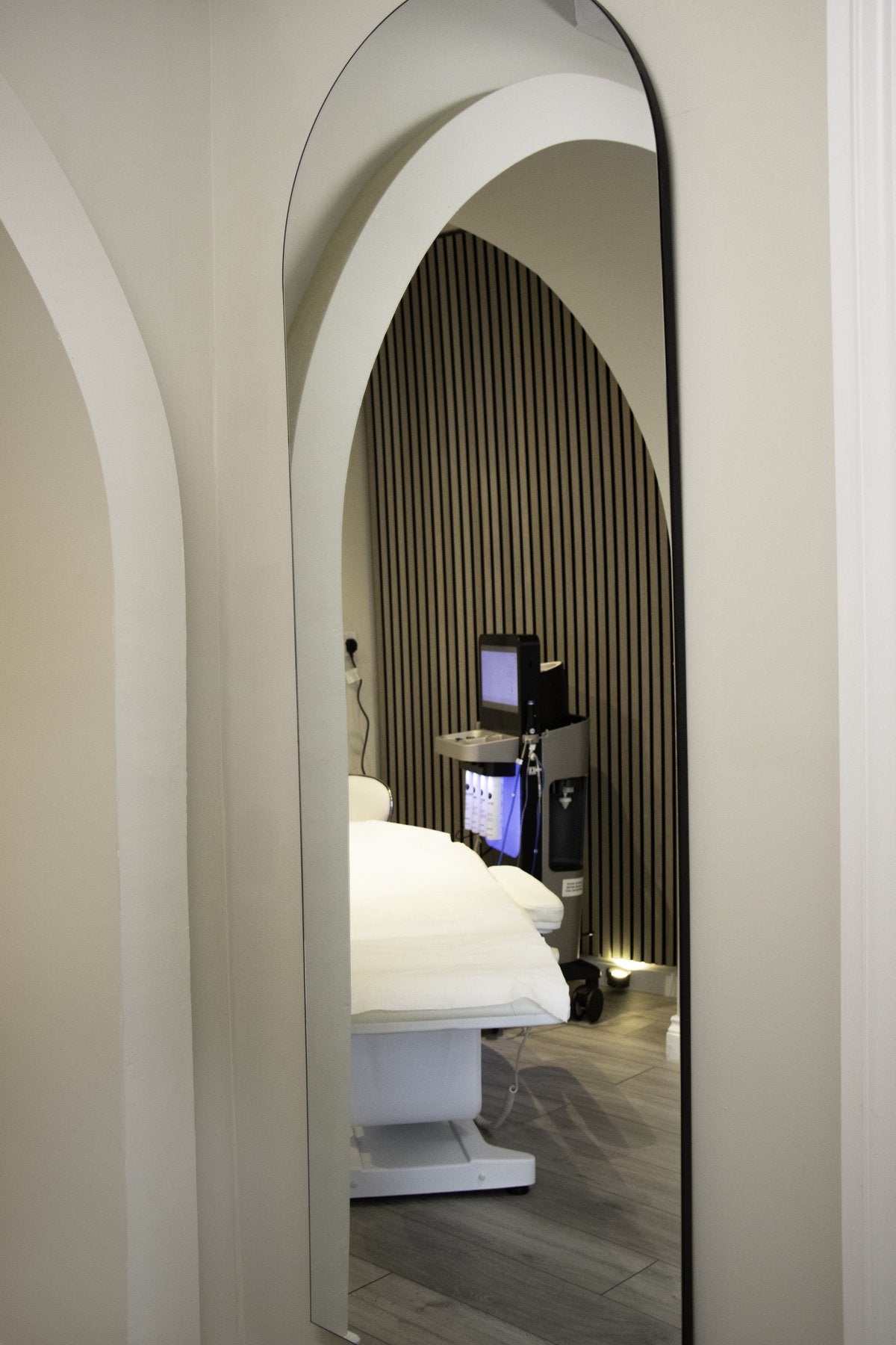 View through an arched mirror into a treatment room at REGEN Clinic Norwich, showing soft lighting, slatted timber feature wall, and a white treatment chair.